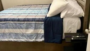 Uma cama de solteiro com colcha listrada e duas almofadas, cabeceira de madeira, mesa de cabeceira pequena com um dispositivo eletrônico preto, e piso coberto por carpete.