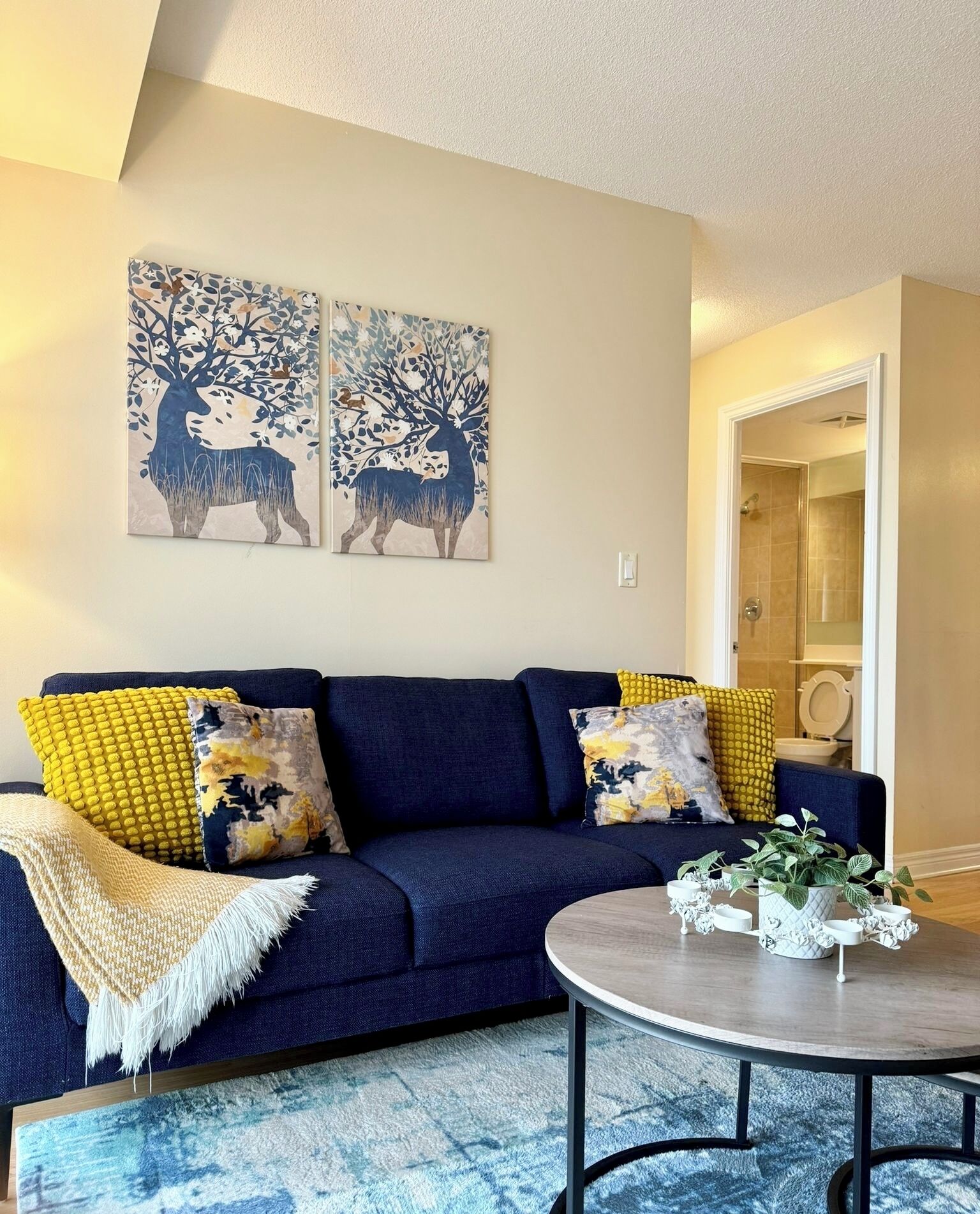 Una sala moderna con un sofá azul oscuro, cojines amarillos y estampados, una mesa redonda de centro con objetos decorativos y dos obras de arte murales que representan ciervos.