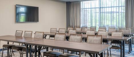 A conference room with multiple tables and chairs, a flat-screen TV mounted on the wall, and large windows with curtains.