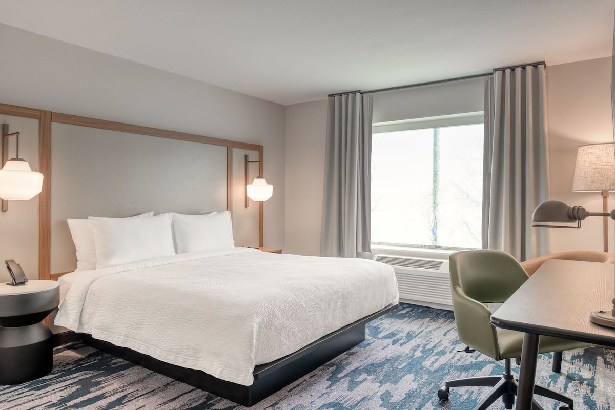 Una habitación de hotel con cama, escritorio, silla, lámpara, teléfono en una mesita de noche y ventana con cortinas.
