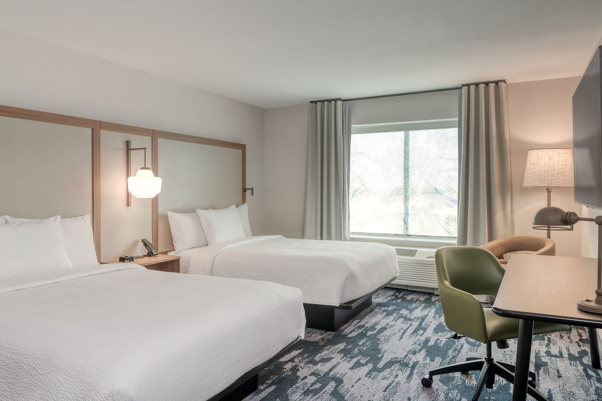 Habitación de hotel con dos camas, un escritorio, una silla, una lámpara y una ventana con cortinas.