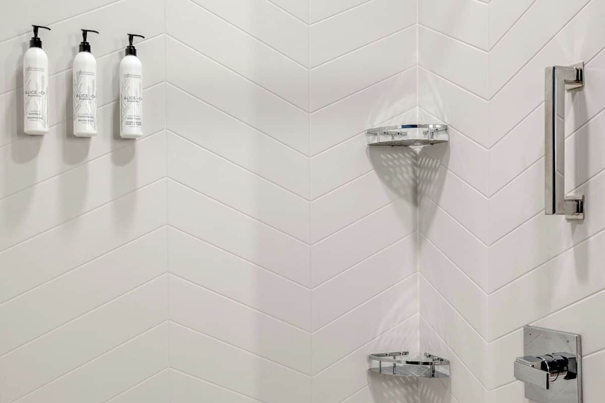 Un baño moderno con pared de baldosas blancas en patrón de espiga, una bañera independiente y tres botellas de artículos de aseo adheridas a la pared.