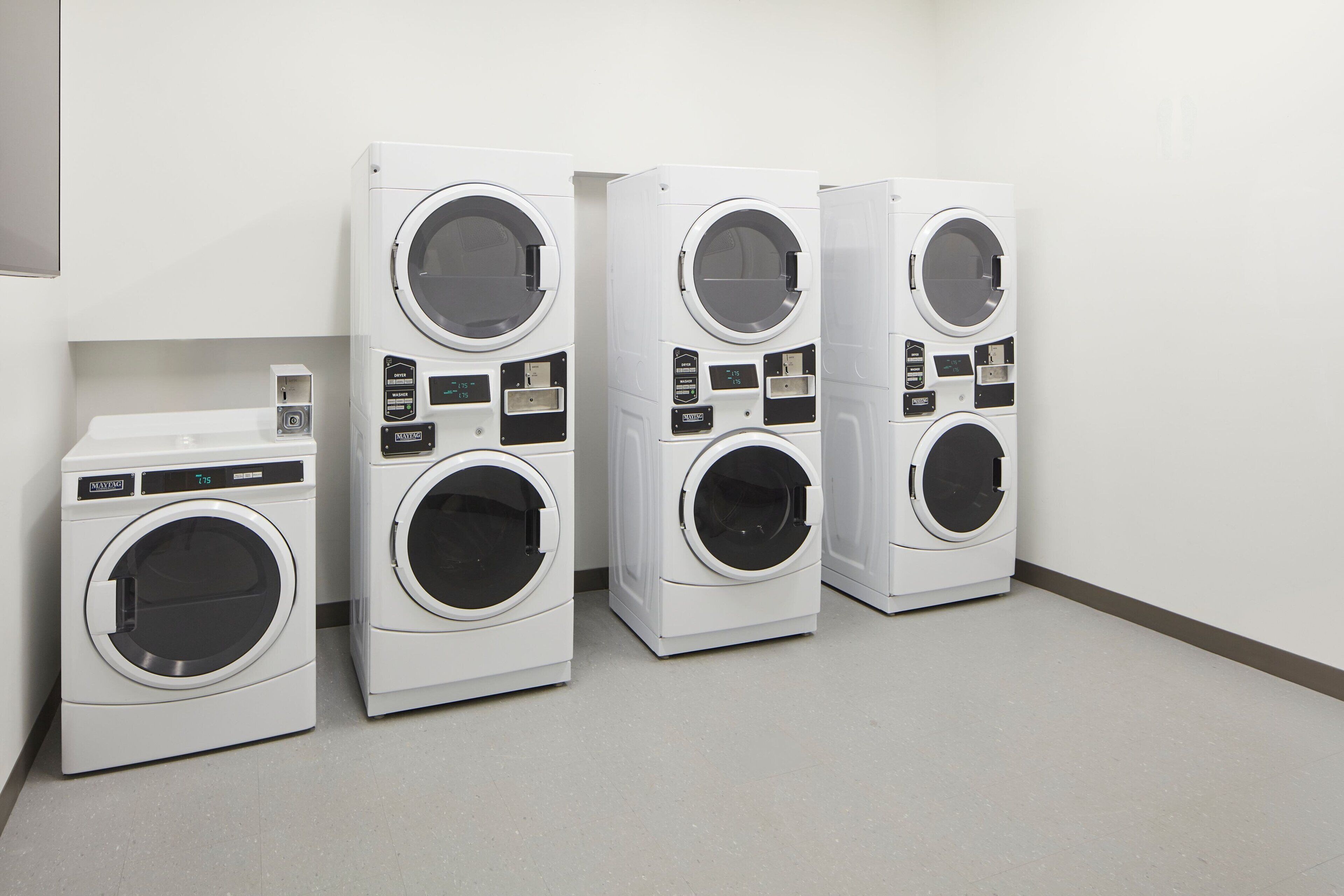 Une rangée de quatre machines à laver à chargement frontal, toutes blanches, se trouve dans une pièce propre et vide, avec un mur blanc uni et un sol de couleur claire.