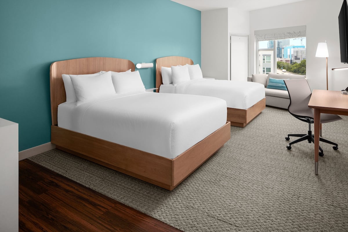 A hotel room with two beds, a desk, a chair, and a view of the city.