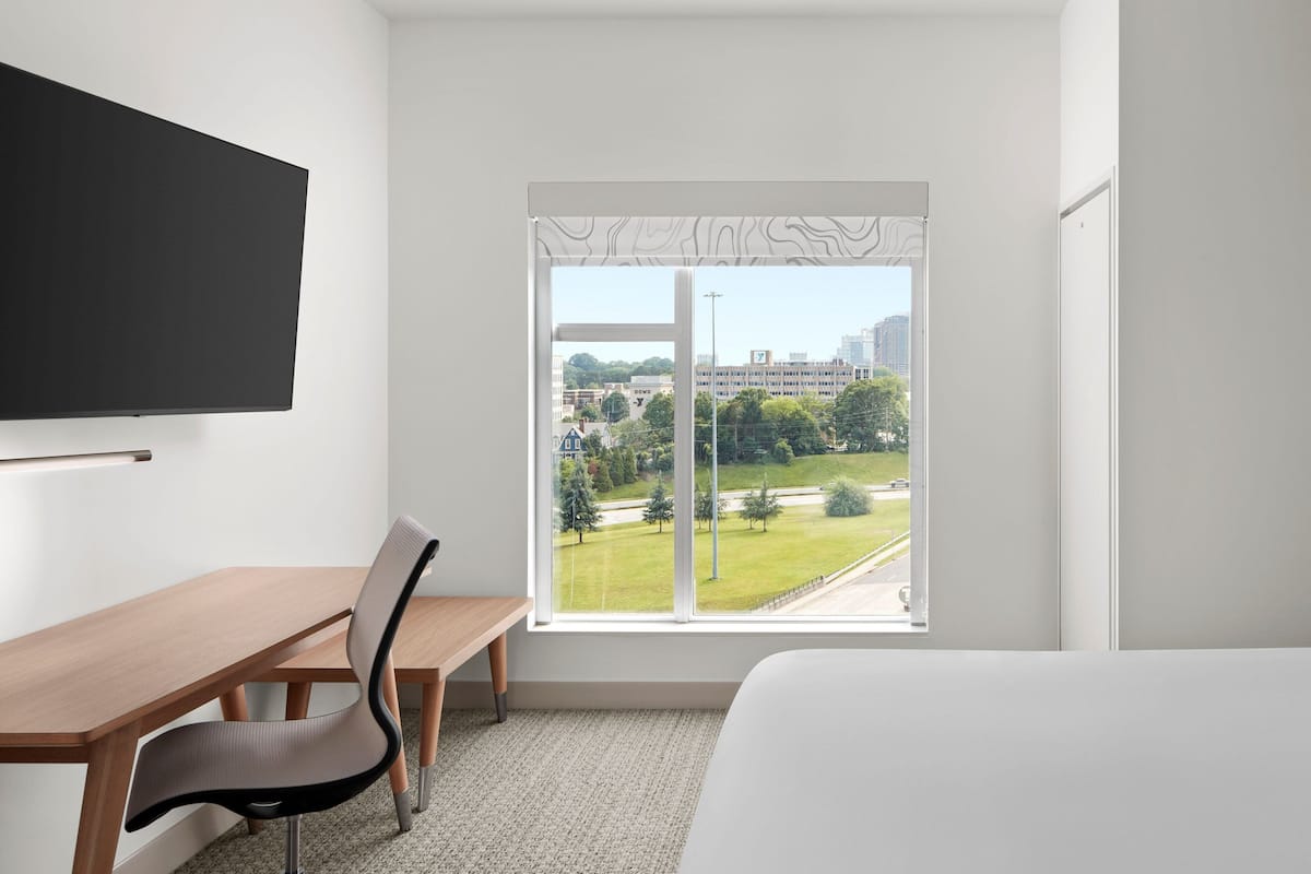 A hotel room with a bed, a desk, and a chair. There is a window with a view of a cityscape and greenery.