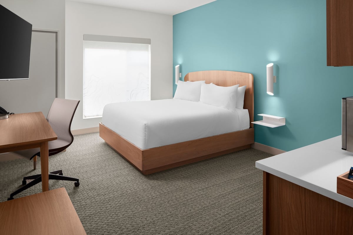 A modern hotel room with a large bed, a desk, a chair, a TV, and a window with a view.