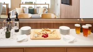 A well-lit kitchen with a wooden countertop, a cutting board with cheese and fruits, wine bottles, glasses, and a dining area with a sofa and table. - Element by Marriott Charlotte Uptown (Charlotte)