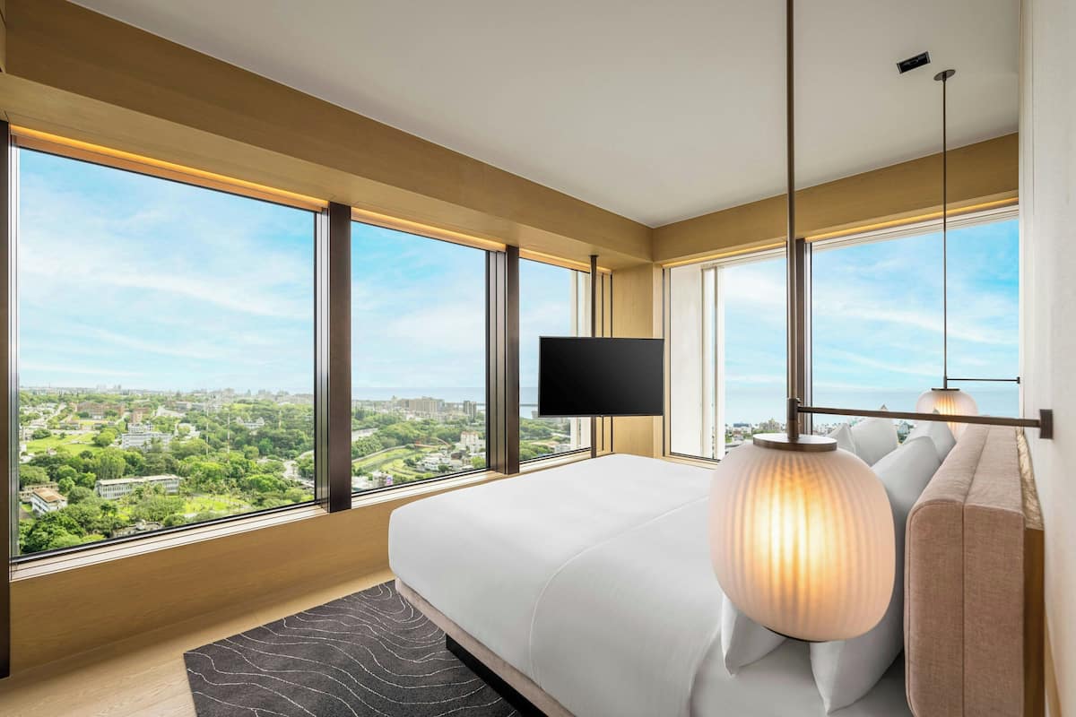 A modern hotel room with a large bed, a flat-screen TV, and a city view through large windows.