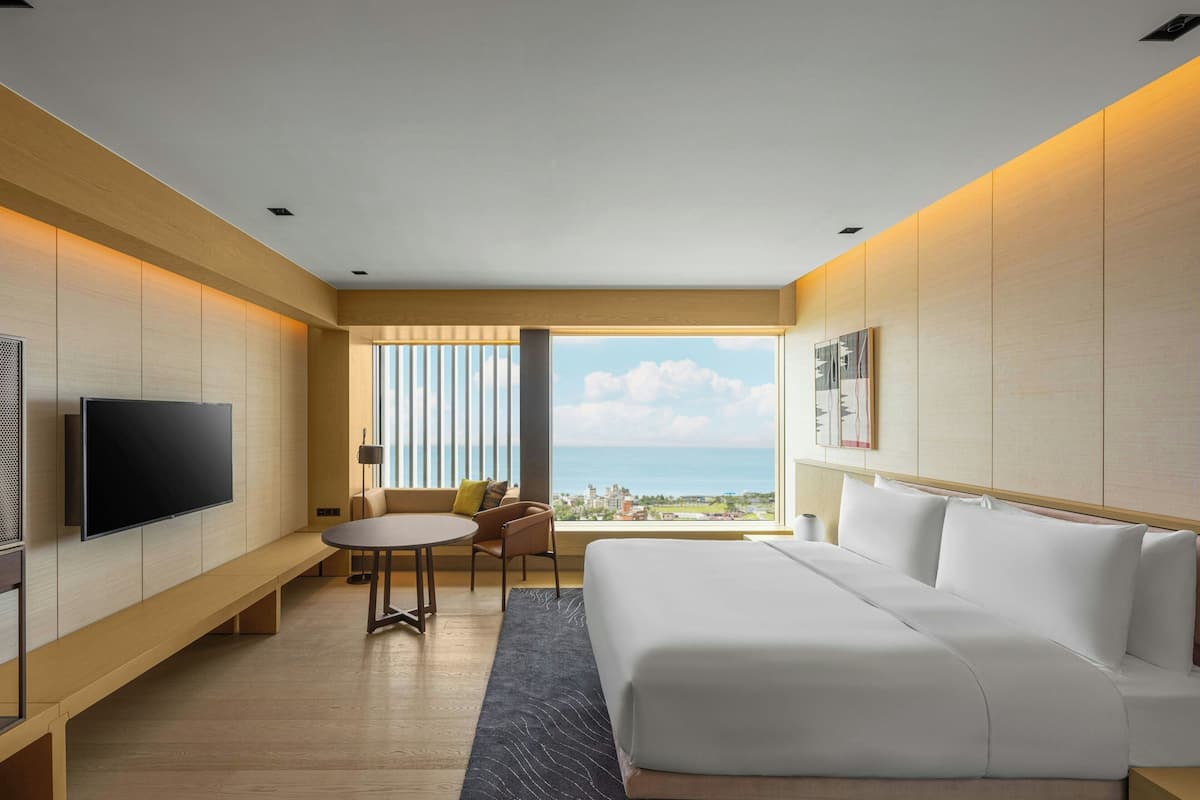 A modern hotel room with a large bed, a flat-screen TV, a seating area with a table and chairs, and a view of the city through large windows.