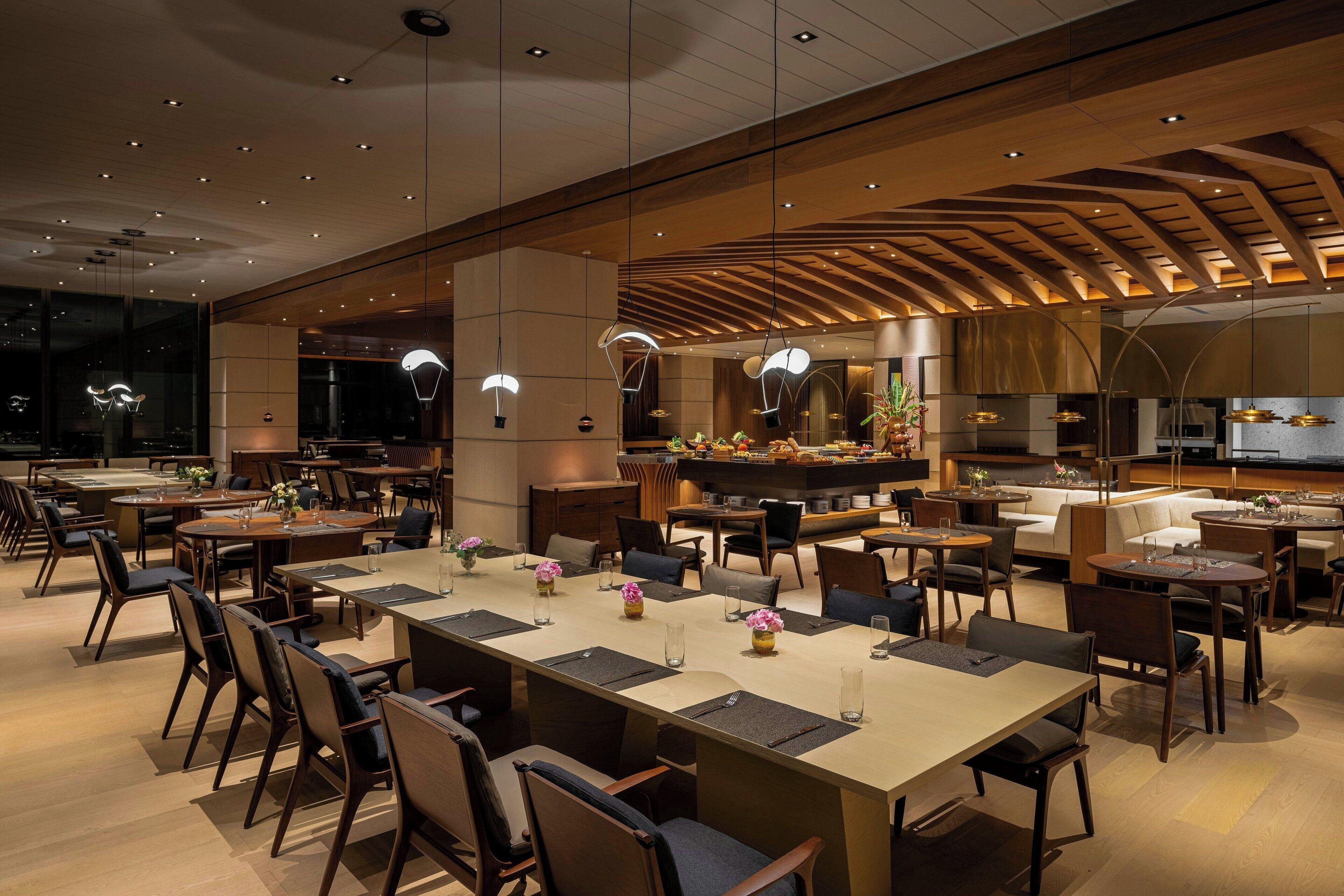 A modern dining area with multiple tables and chairs, a central kitchen area, and a high ceiling with wooden beams.