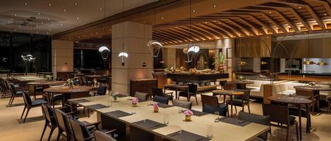 A modern dining area with multiple tables and chairs, a central kitchen area, and a high ceiling with wooden beams.