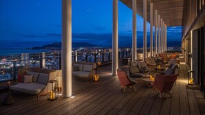 Un lounge en la azotea con varias zonas de estar, grandes pilares y vistas a la ciudad al anochecer.