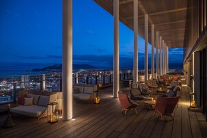 Un lounge en la azotea con varias zonas de estar, grandes pilares y vistas a la ciudad al anochecer.
