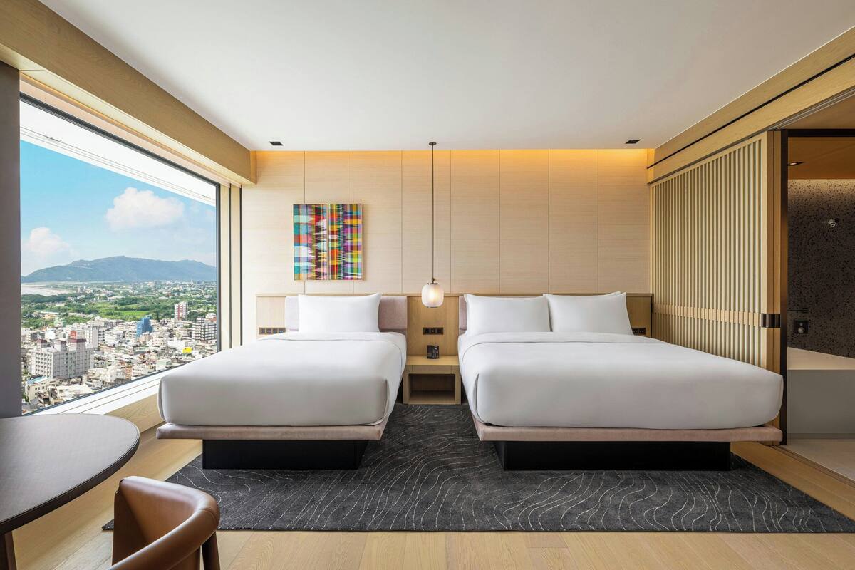 A modern hotel room with two beds, a city view, a small table, and a chair.