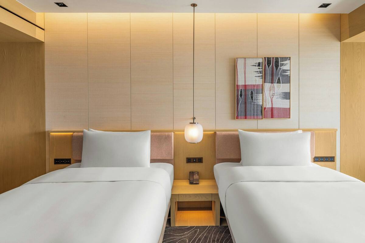 A hotel room with two beds, a bedside table with a lamp, a phone, and a wall-mounted artwork.