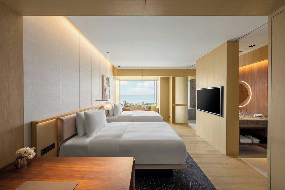 A modern hotel room with two beds, a TV, a desk, and a bathroom with a vanity and mirror.