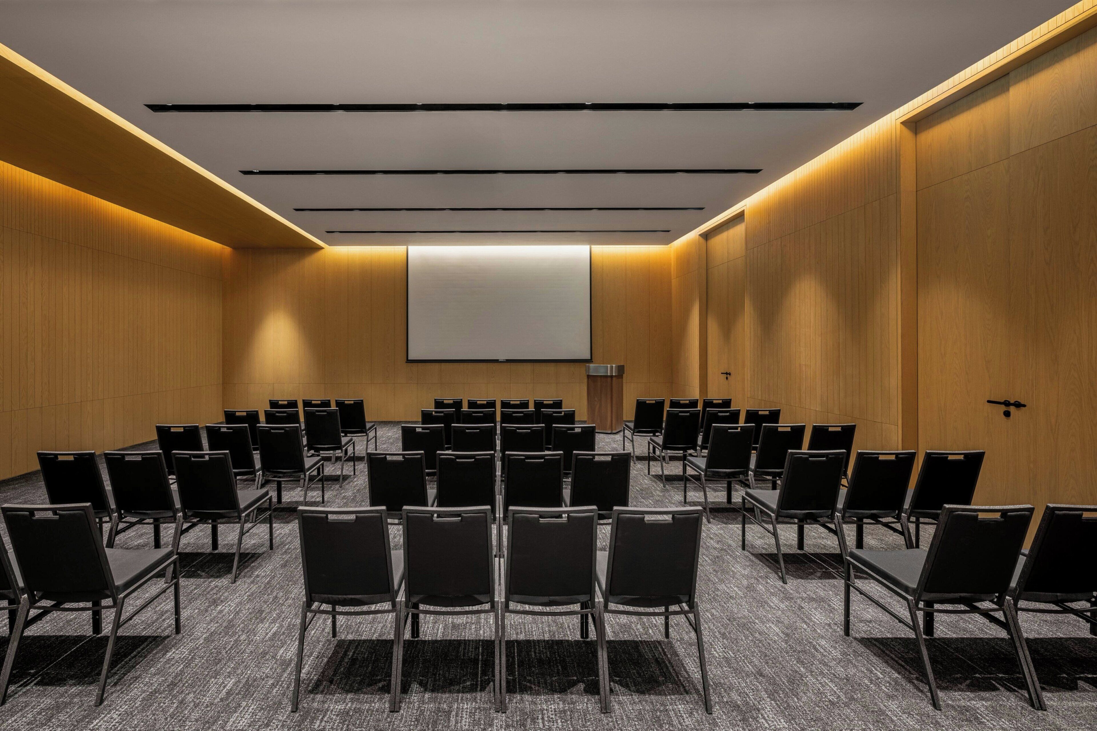 A conference room with rows of chairs, a large screen, and wooden paneling.