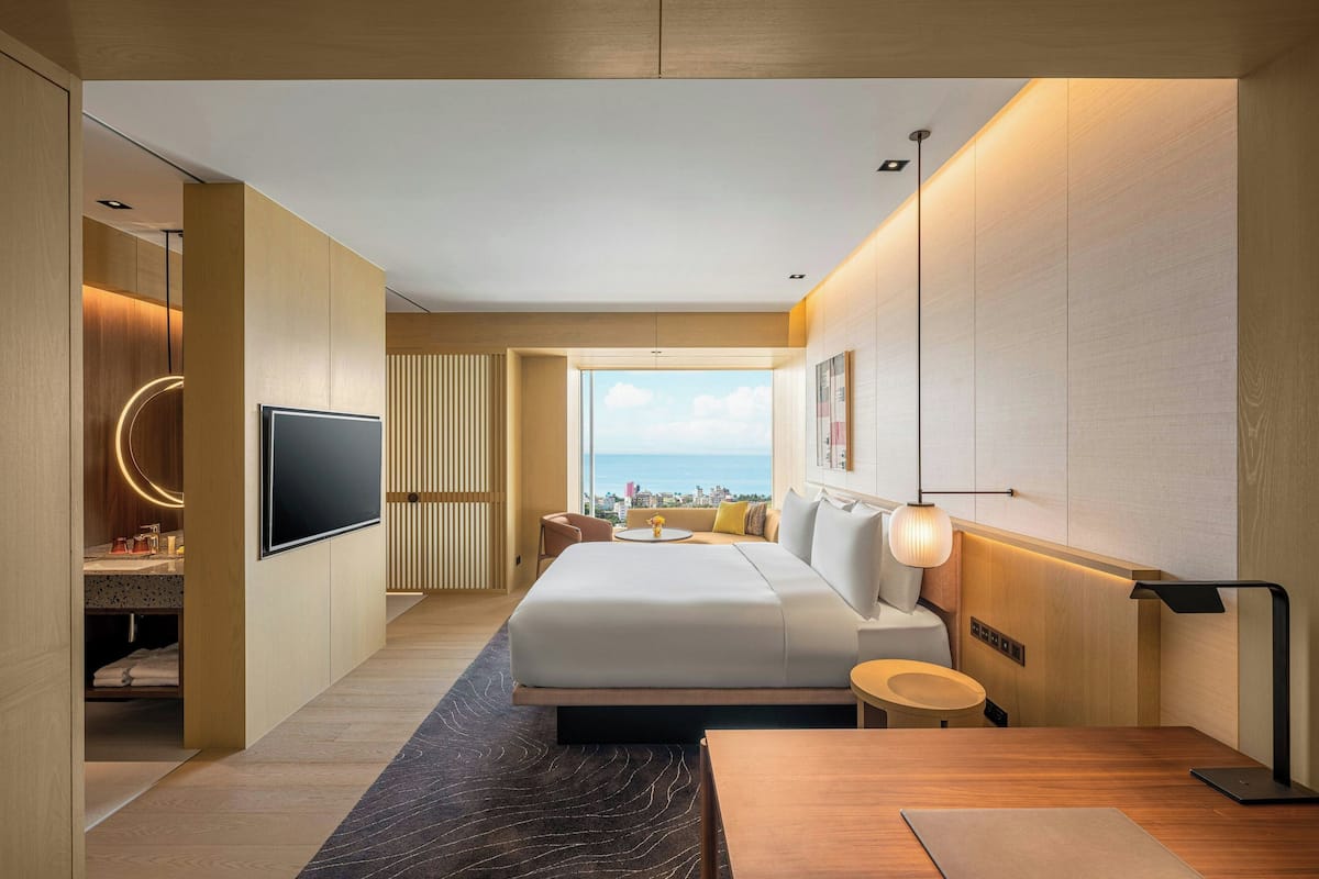 A modern hotel room with a large bed, a TV, a desk, and a view of the city.