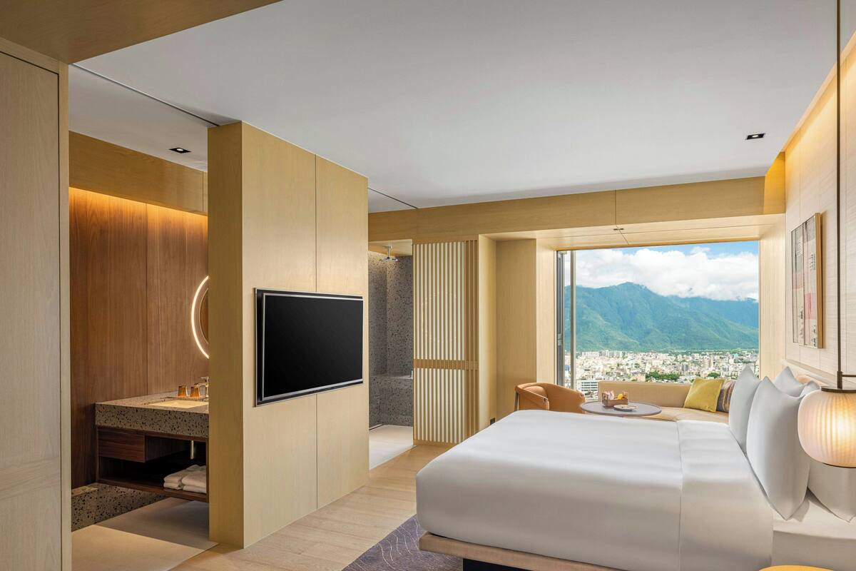 A modern hotel room with a large bed, a TV, a bathroom with a vanity, and a view of mountains.