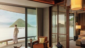 Une chambre d’hôtel moderne dotée d’une grande fenêtre donnant sur une étendue d’eau et une île lointaine. On y trouve un coin salon avec un canapé et des fauteuils, ainsi qu’une petite table agrémentée d’un objet décoratif.
