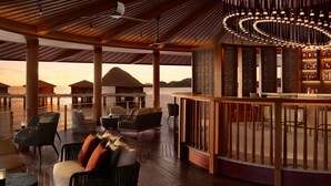 Une terrasse en bois dotée de sièges et d’un espace bar, offrant une vue sur une étendue d’eau, avec des bungalows sur pilotis en arrière-plan.