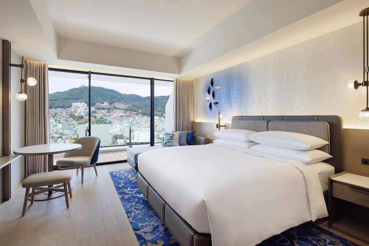 Een moderne hotelkamer met een groot bed, een kleintje tafeltje, een stoel en uitzicht op een stadsgezicht.