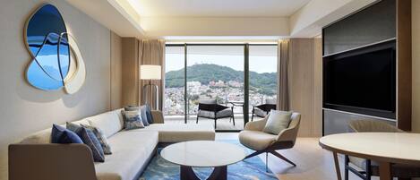 A modern hotel room with a large window, a sofa, a round coffee table, and a TV.