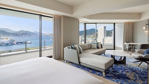 Une chambre d’hôtel moderne dotée d’un grand lit, d’un canapé, d’une table à manger et offrant une vue sur la mer et le paysage urbain.