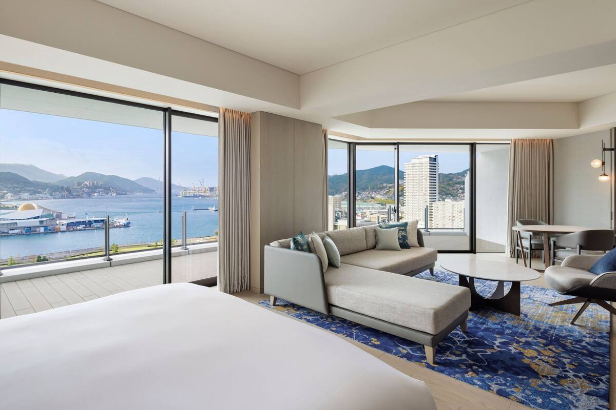 Een moderne hotelkamer met een groot bed, een bank, een eettafel en uitzicht op zee en de stad.