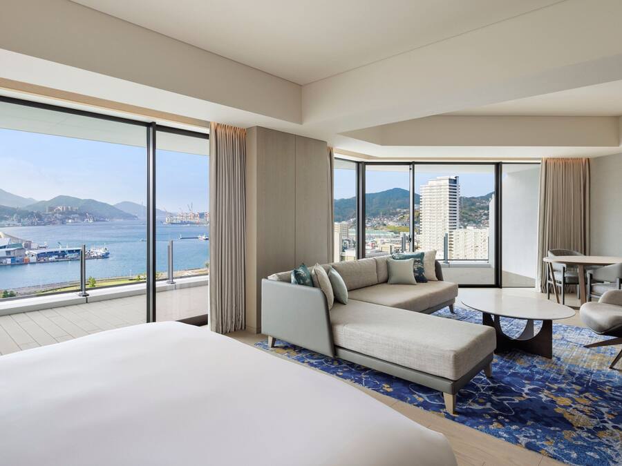 A modern hotel room with a large bed, a sofa, a dining table, and a view of the sea and cityscape.