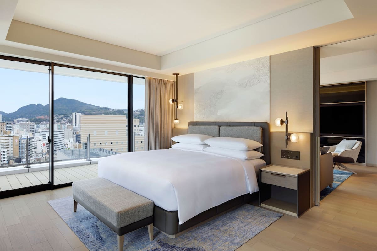 Een moderne hotelkamer met een groot bed, nachtkastje, een bankje, een televisie en uitzicht over de stad.