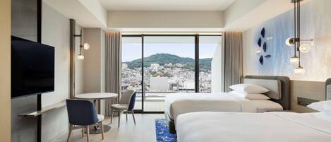 A hotel room with two beds, a TV, a small table, and a chair. The room has a large window with a city view.