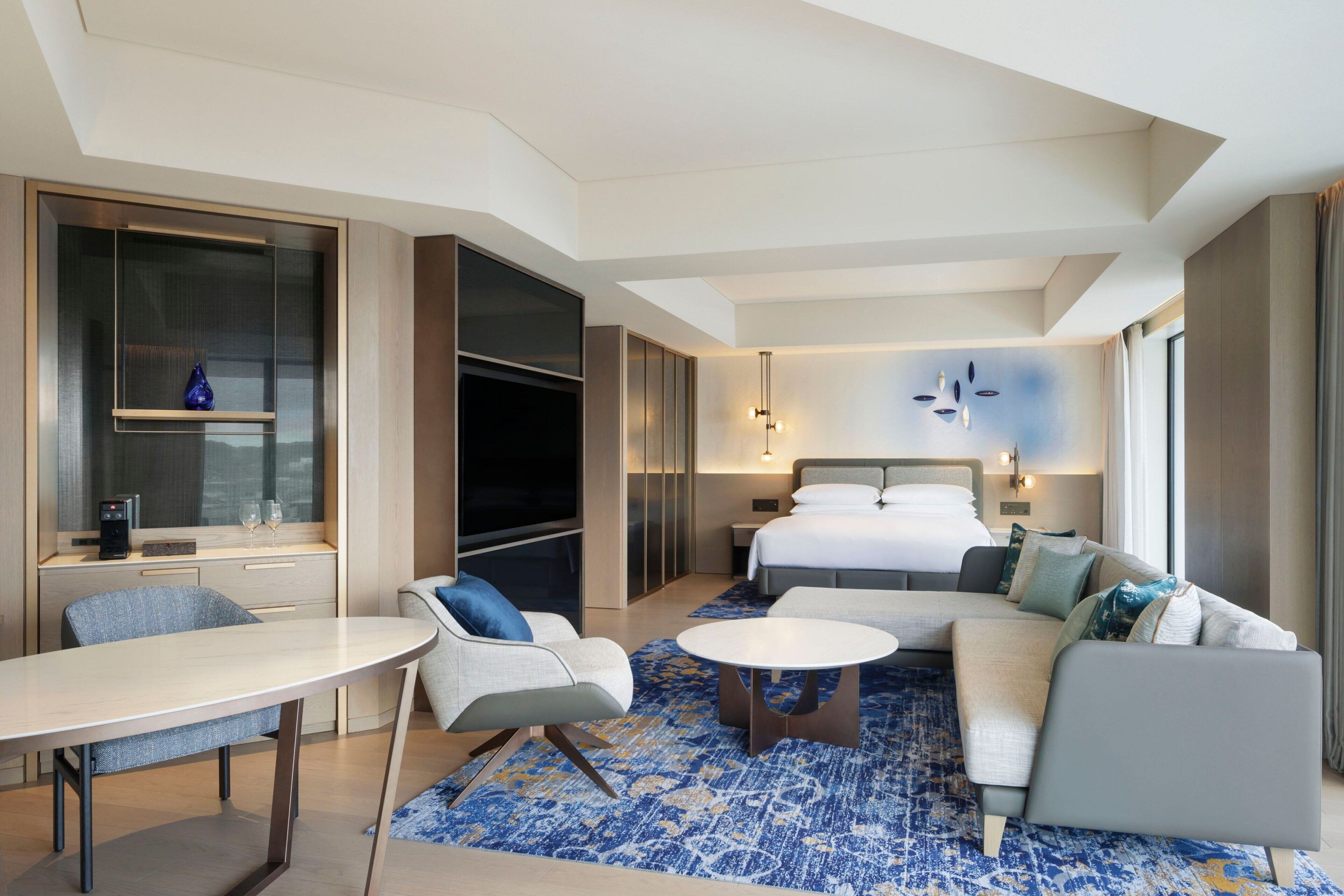 A modern hotel room with a bed, sofa, dining table, and a TV.