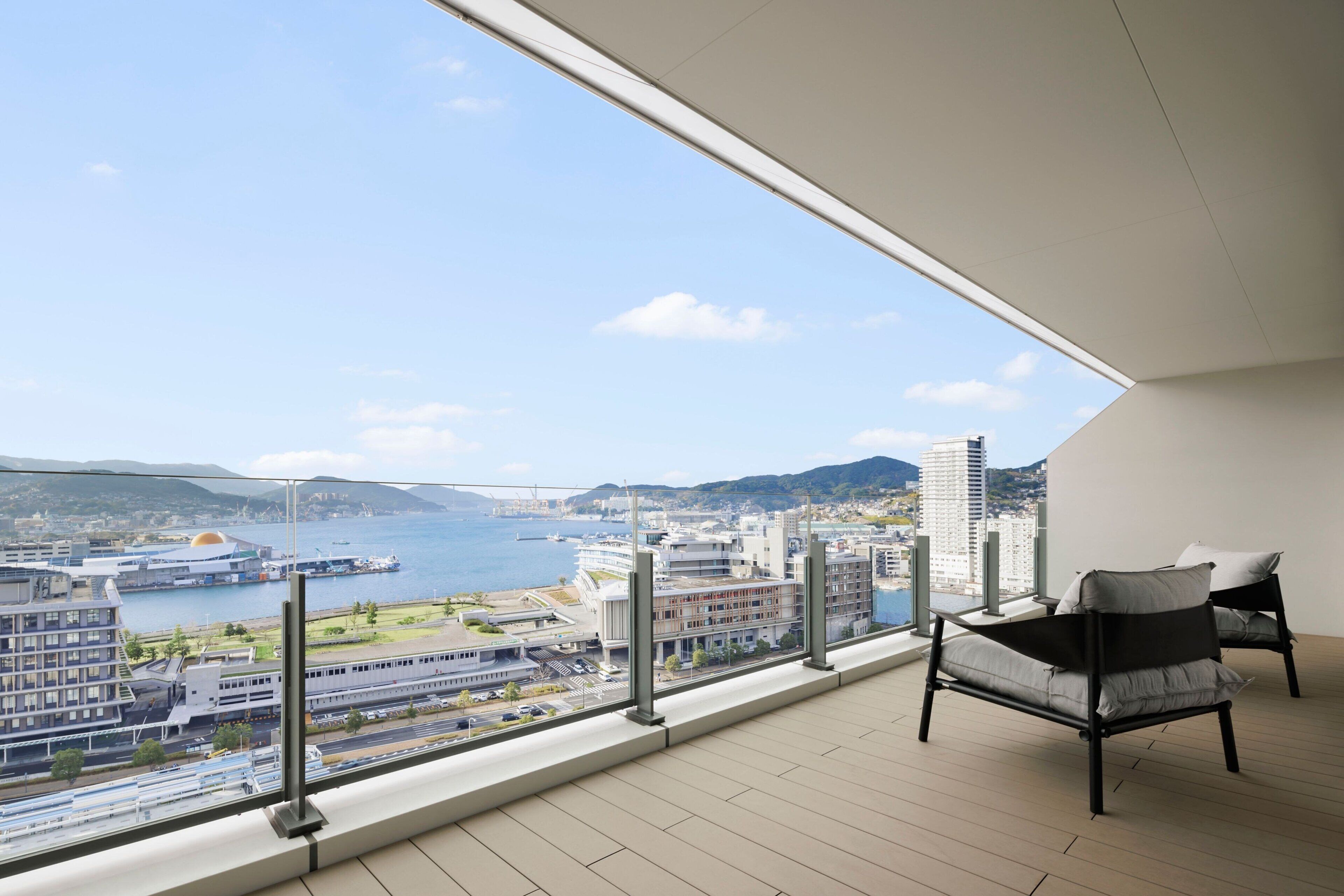 A balcony with a view of a cityscape, including a bridge and buildings. There are two chairs and a table on the balcony.