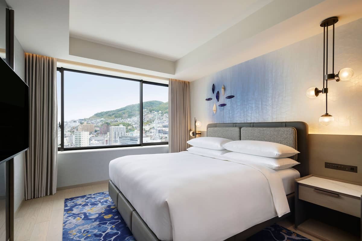 Hotelkamer met een groot bed, een televisie, een nachtkastje en uitzicht op een stadsgezicht door een groot raam.