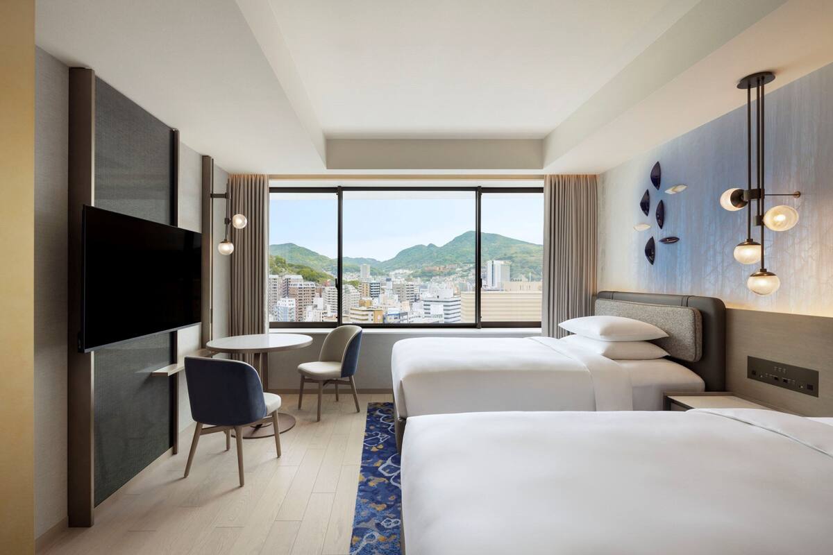 Een hotelkamer met twee bedden, een televisie, een klein tafeltje met een stoel en een groot raam met uitzicht op de stad.