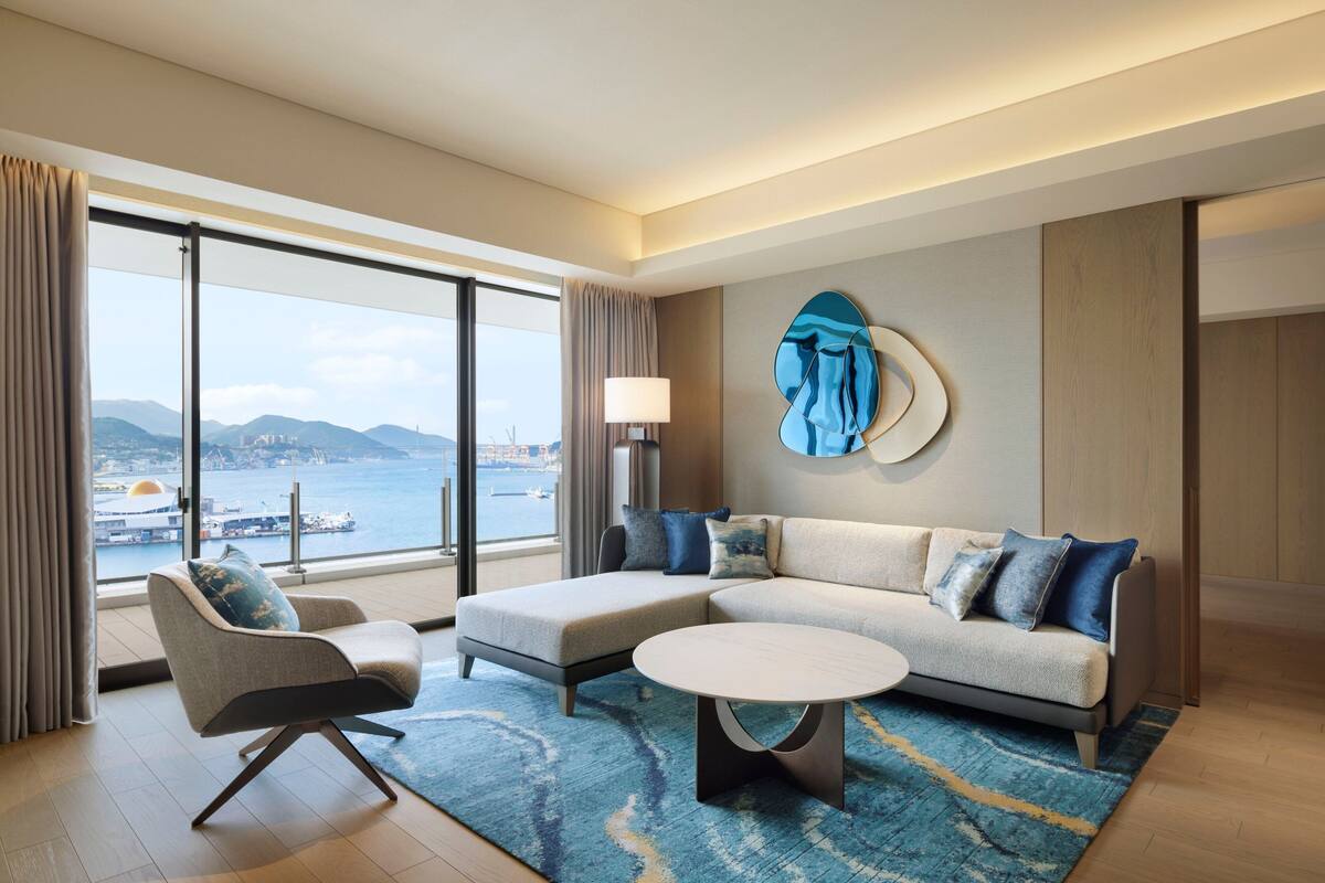 Een moderne hotelkamer met een groot raam met uitzicht op zee en bergen. Er is een bank, een stoel en een ronde salontafel.
