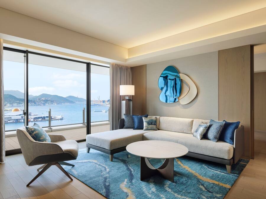 A modern hotel room with a large window offering a view of the sea and mountains. It includes a sofa, a chair, and a round coffee table.