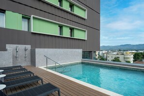 Una piscina en la azotea con cubierta de madera, barandilla de vidrio y vista panorámica de la ciudad.