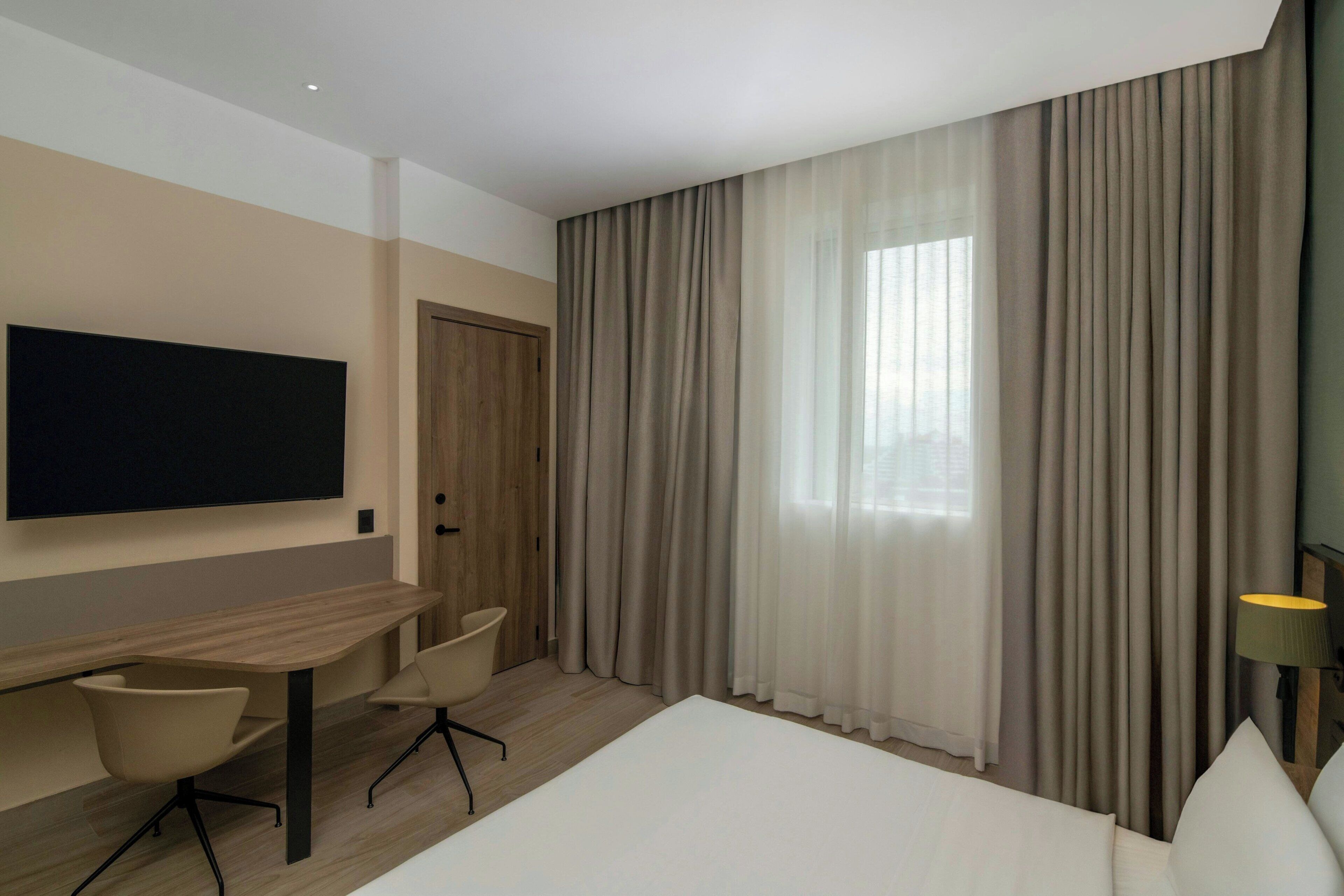 A modern hotel room with a large bed, a desk with two chairs, a flat-screen TV, and large windows with curtains.