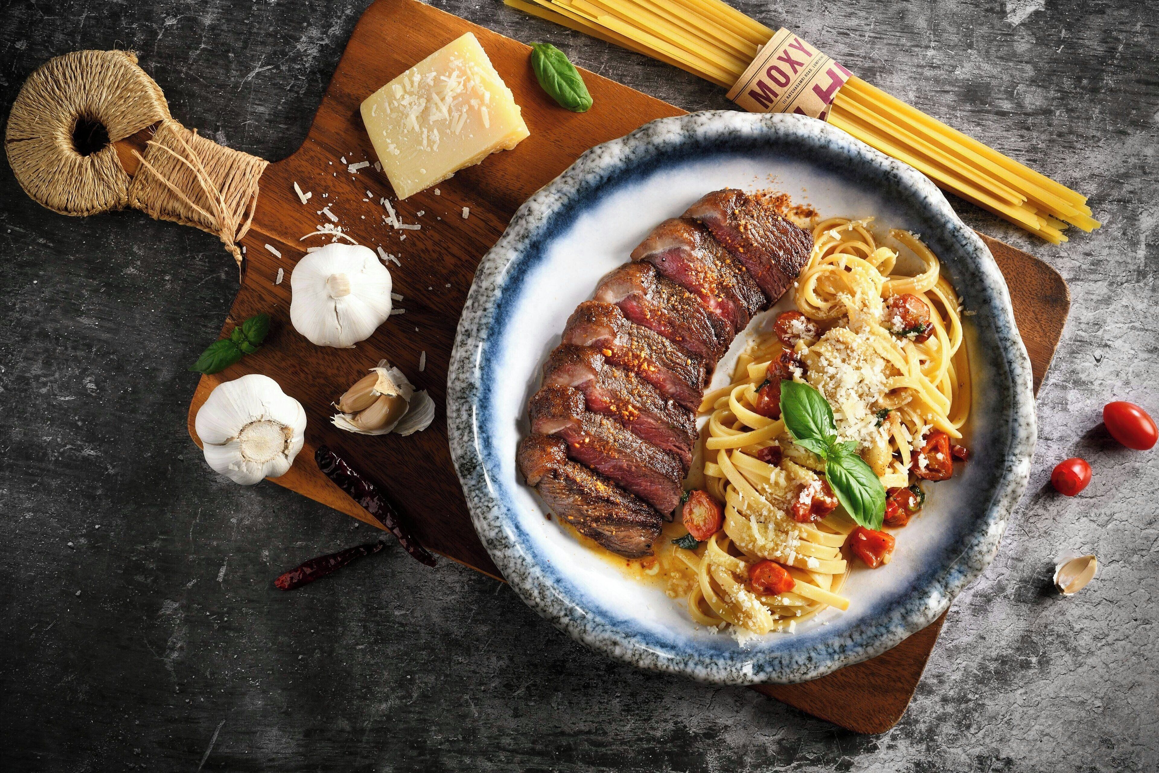 Un plato de pasta con carne y queso, rodeado de ajo, tomates y albahaca sobre una superficie oscura.