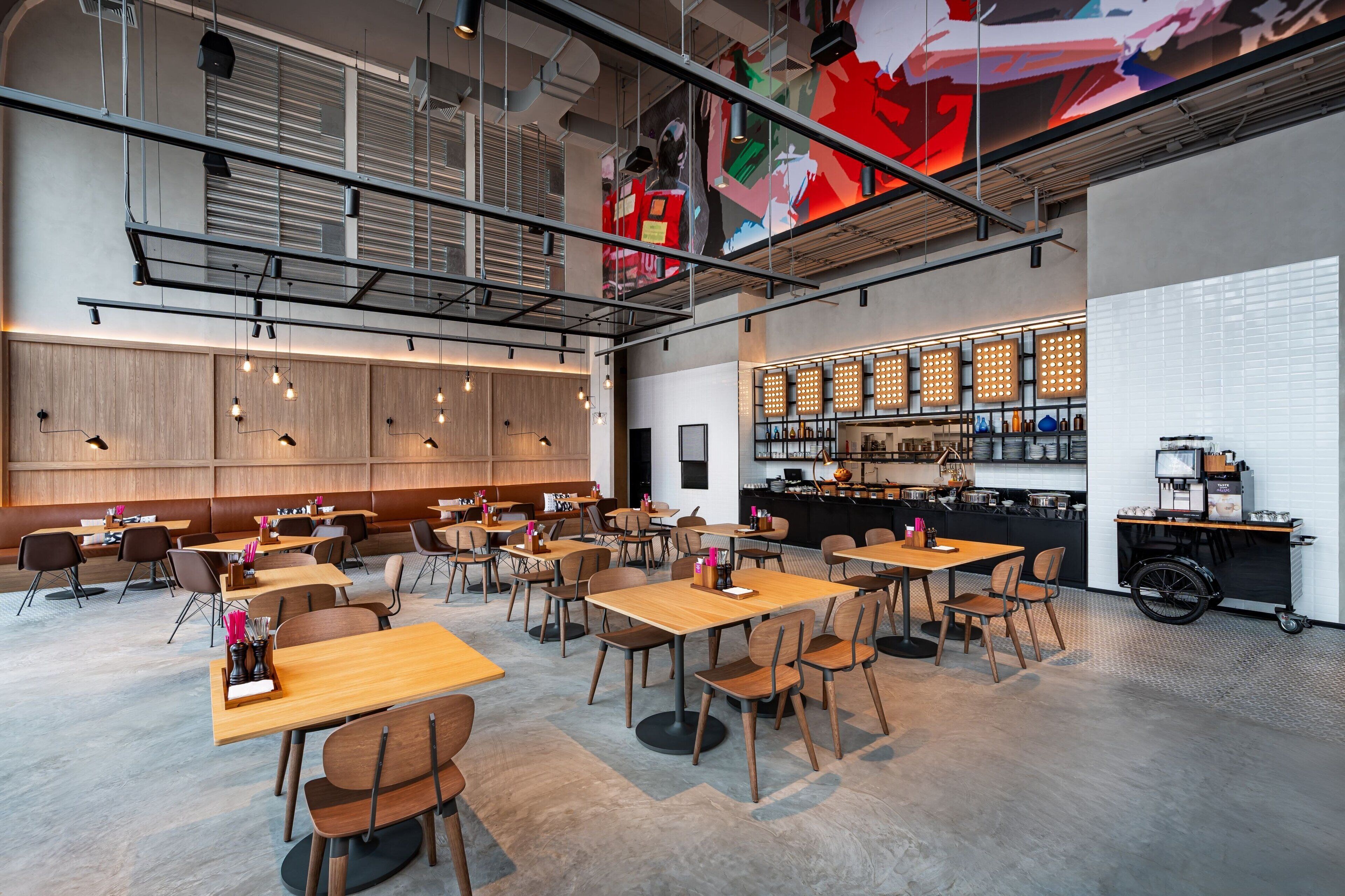 Ein modernes Restaurant mit hohen Decken, freiliegenden Lüftungsrohren und einer Bar mit einer Flaschenauslage. Tische sind großzügig angeordnet und mit Stühlen bestückt.