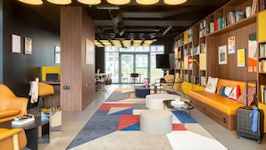 A modern hotel lobby with a central seating area, a bookshelf, and a large window providing a view of the outdoors. - Moxy Paris Clamart (Clamart)