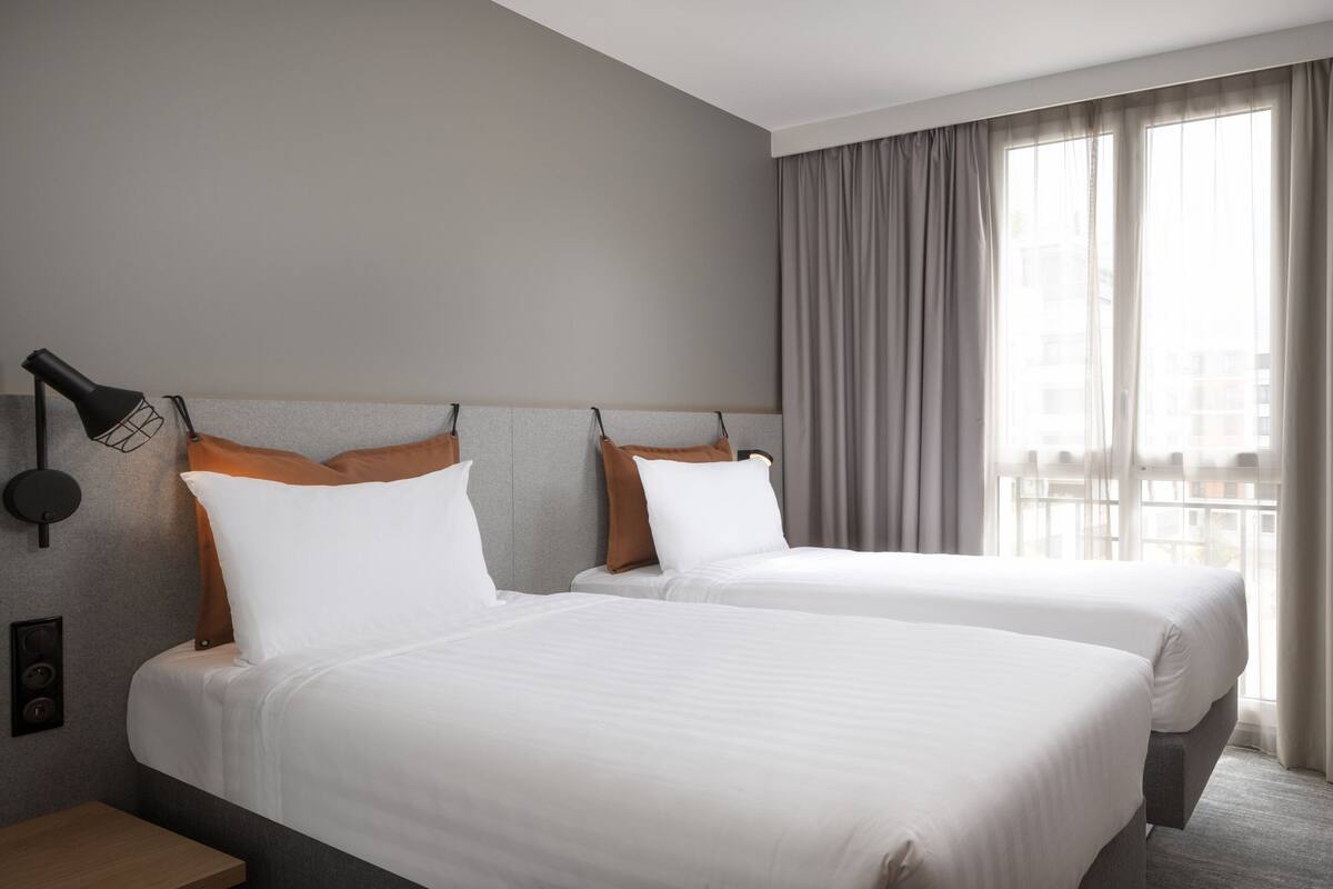Quarto de hotel com duas camas, mesa de cabeceira, abajur e vista da cidade por grandes janelas.