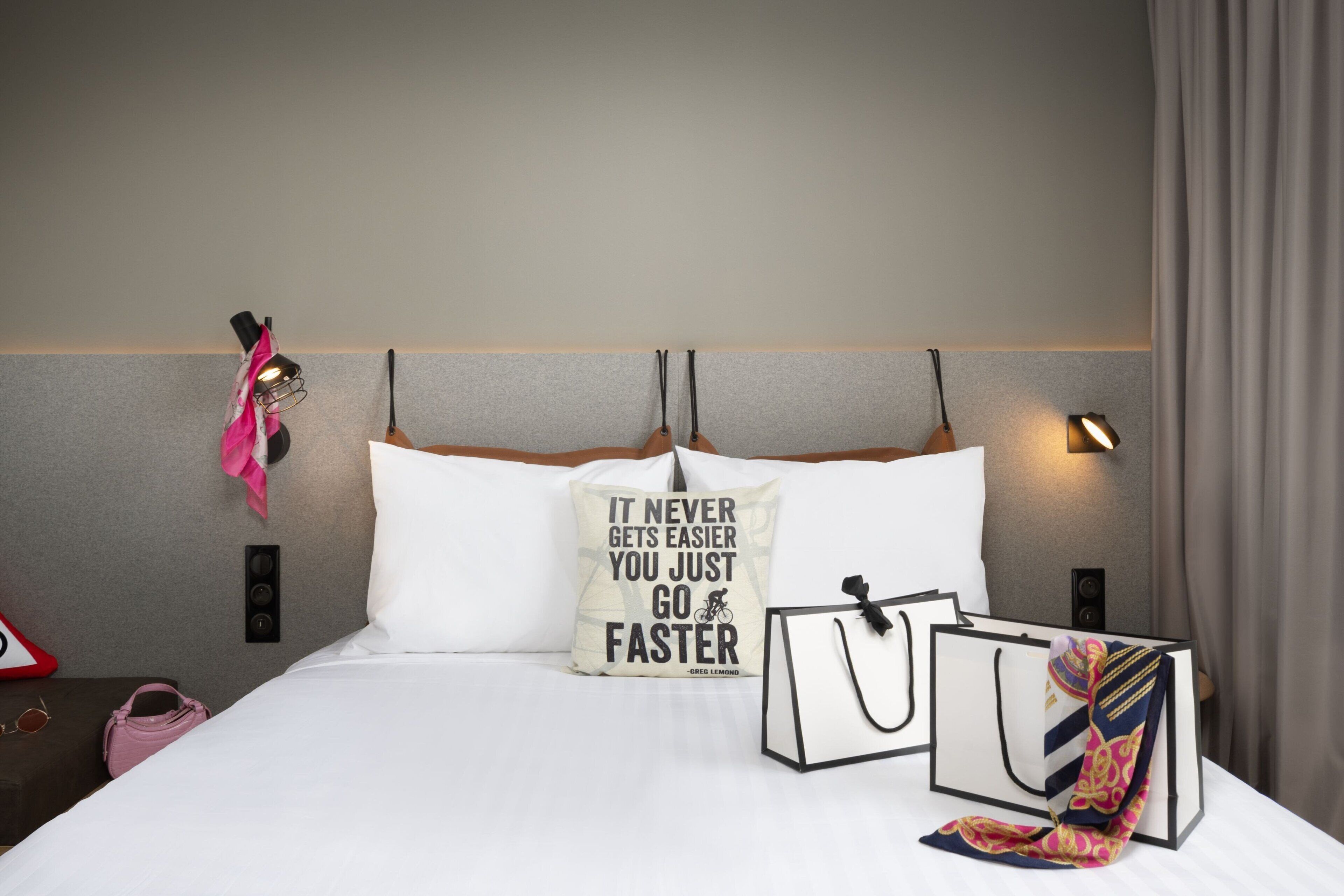 Un lit bien fait, agrémenté d’un coussin aux motifs inspirants, une table de chevet avec un sac à main et des lunettes de soleil, et une lampe fixée au mur.