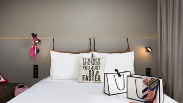 Un lit bien fait, agrémenté d’un coussin aux motifs inspirants, une table de chevet avec un sac à main et des lunettes de soleil, et une lampe fixée au mur.
