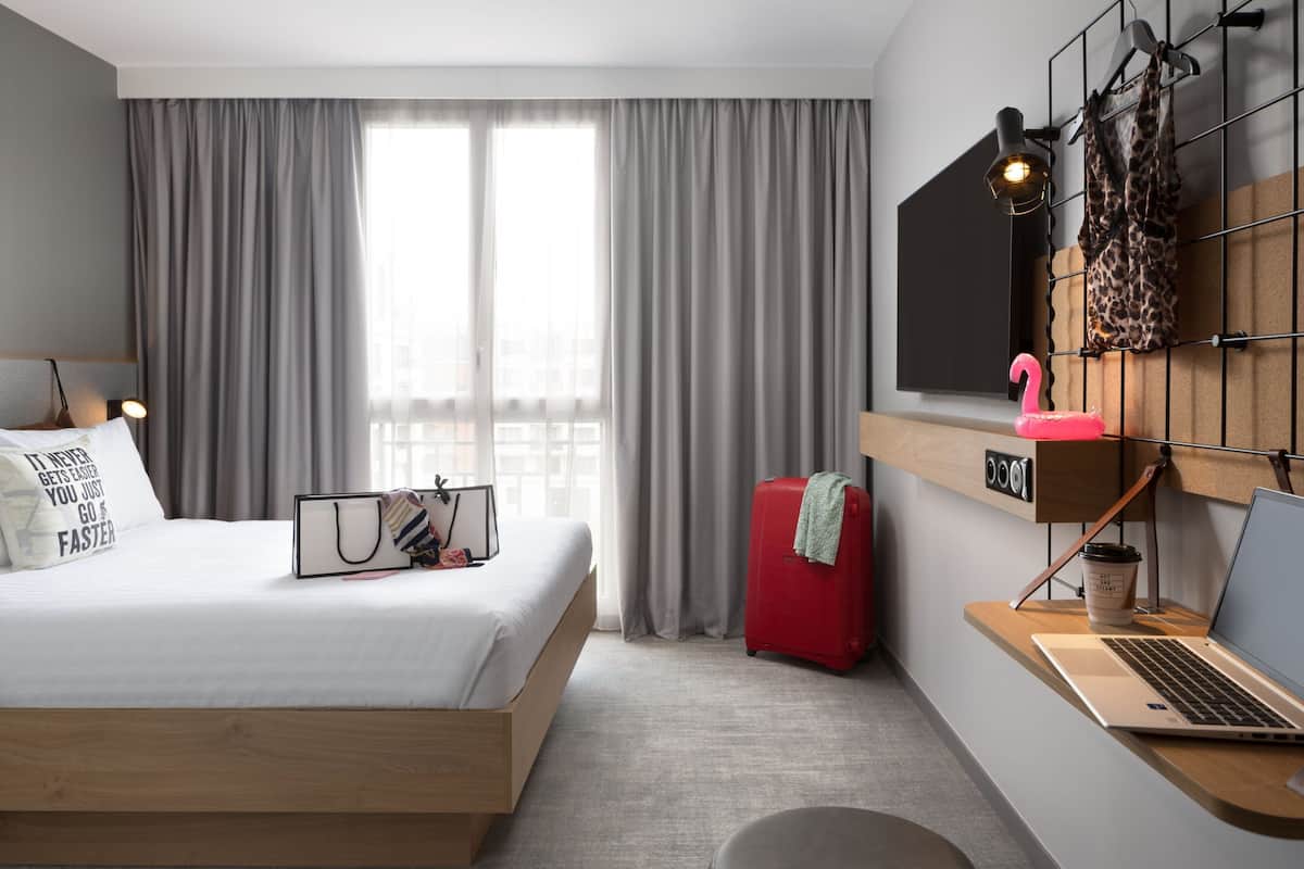 Quarto de hotel moderno com cama, escrivaninha, laptop, mala e janela com cortinas.