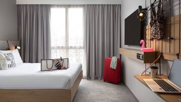 A modern hotel room with a bed, a desk with a laptop, a suitcase, and a window with curtains.