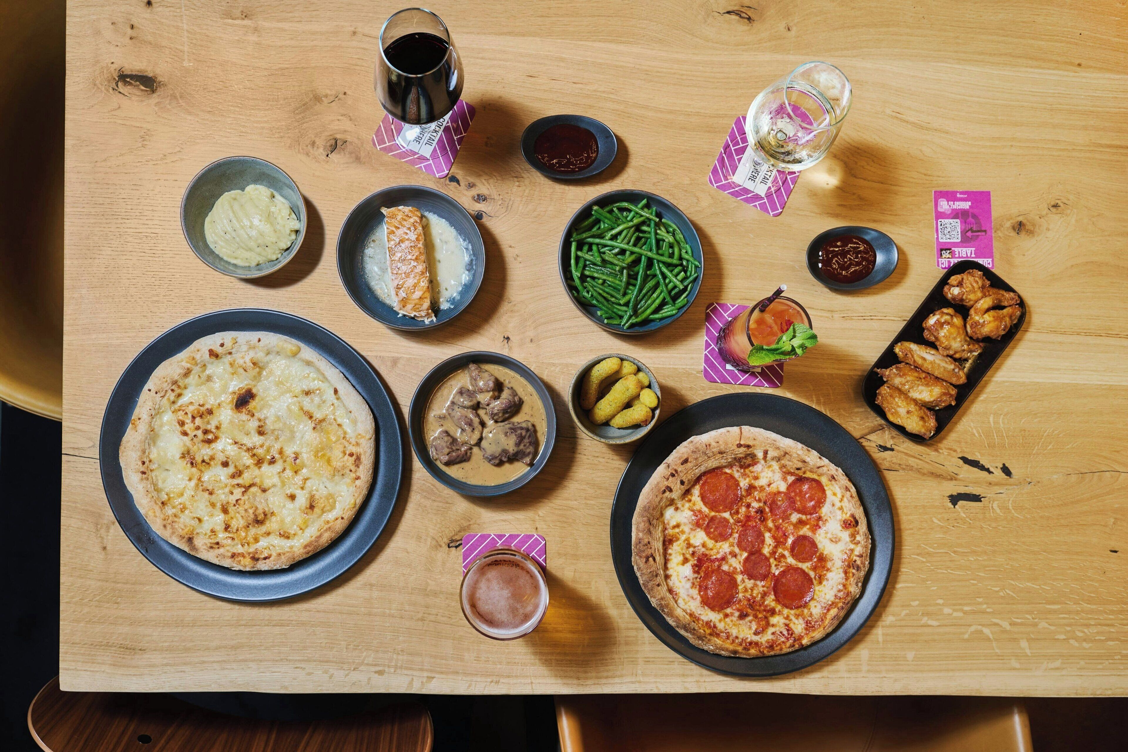 Ett träbord med olika rätter, inklusive pizza, gröna bönor och en tallrik med kycklingvingar.