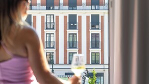 A woman in a pink dress standing by a window, holding a glass of wine. The window overlooks a building with multiple balconies and a dome structure. - Moxy Paris Clamart (Clamart)